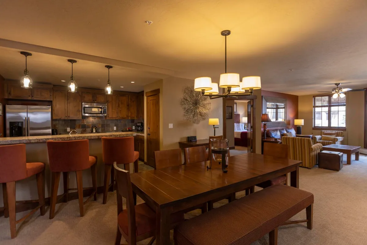 The image showcases a spacious, open-concept living area featuring a stylish dining table, inviting bar stools at a granite-topped kitchen island, and plush seating in the living room. Warm lighting and tasteful decor create a cozy, communal atmosphere ideal for gathering, sharing meals, and relaxing after a day on the slopes.