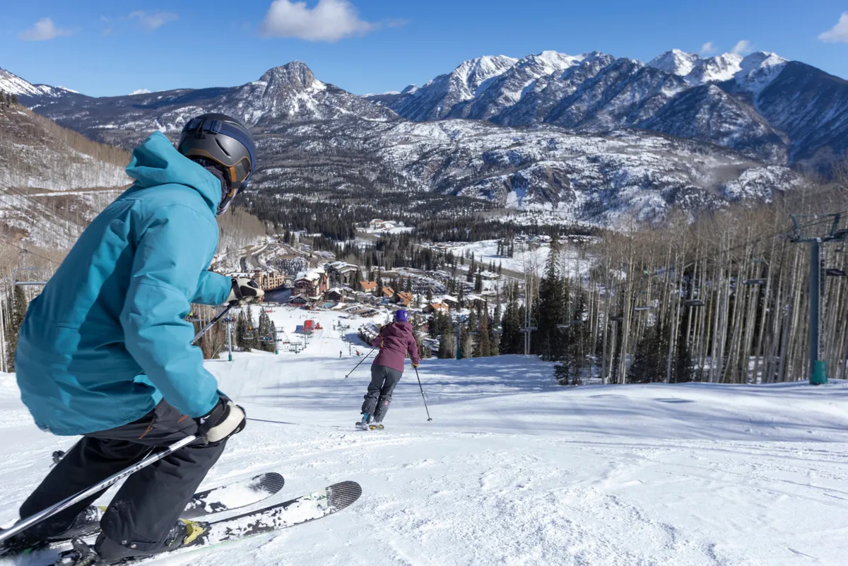 An exterior shot showing skiers descending a snow-covered mountain slope with the vibrant ski village nestled below. The dramatic mountain backdrop and dynamic skiing action convey the thrill of easy access to lifts and trails.