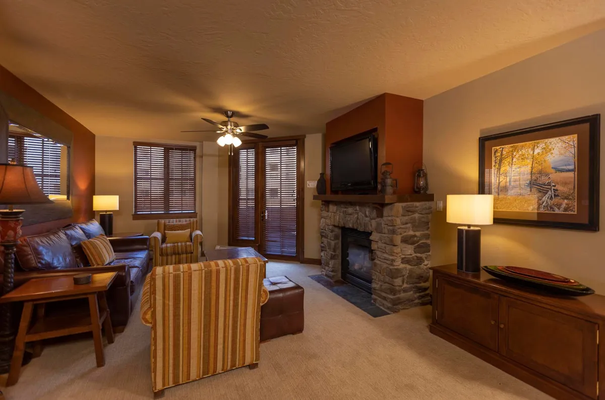 A warm and welcoming living area featuring a stunning stone fireplace, plush leather sofa, striped accent chair, and tasteful artwork. Large windows (not shown but implied by lighting) suggest a seamless connection to the outdoors, perfect for relaxing after a day of mountain adventures.