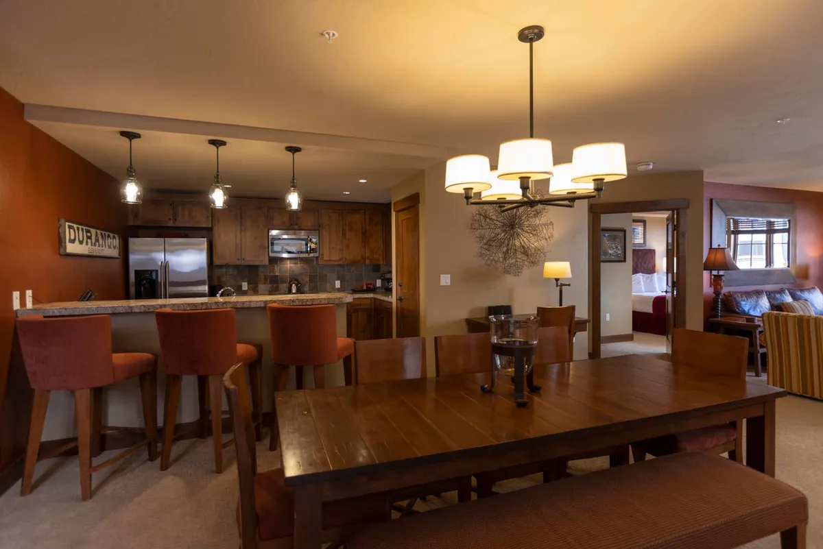 A spacious, inviting dining area complete with an elegant chandelier and a large table set for meals. The kitchen island and bar stools connect the dining area to the gourmet kitchen, while a cozy sitting area is visible in the background. The open layout encourages both intimate family dinners and lively celebrations.
