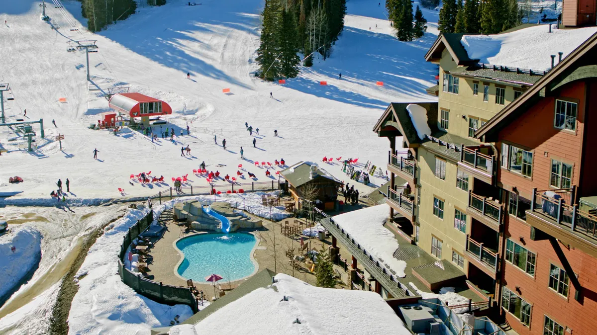 A sweeping view of the snowy lodge grounds featuring ski slopes, a sparkling swimming pool, multiple lodge buildings, and social groups enjoying the vibrant winter scene. The festive energy and diversity of activities are front and center.