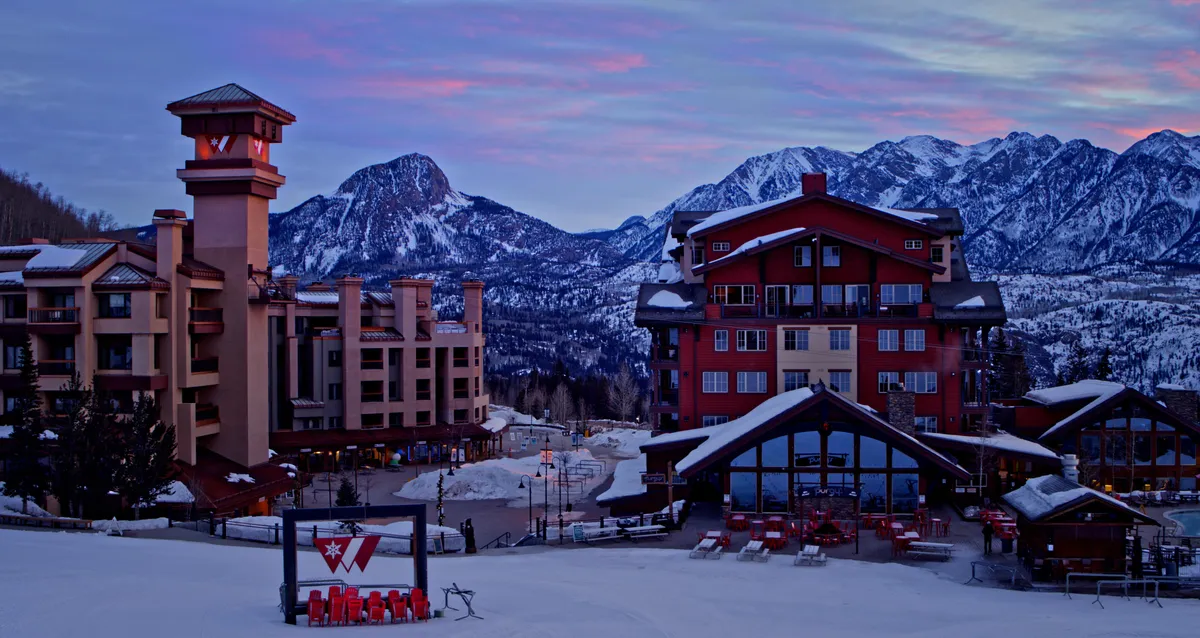 A cozy exterior scene featuring the lodge’s architecture against a dramatic mountain backdrop, with snow on the ground and an inviting outdoor seating area. The setting suggests après-ski relaxation and opportunities to take in stunning views or gather by a fire pit.