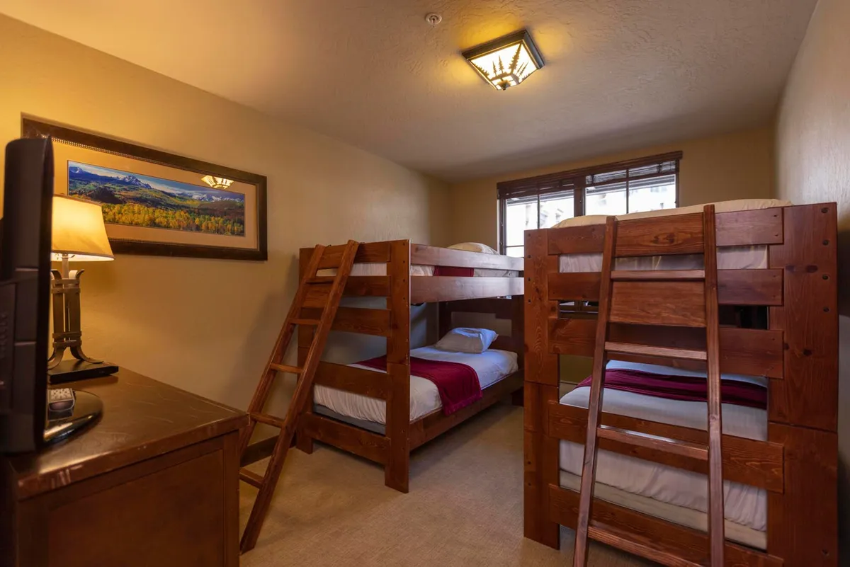 A cozy bunk room with wooden bunk beds, a lamp for reading, framed artwork, and a window illuminating the space. The inviting setup is ideal for children, featuring playful yet relaxing touches that encourage late-night chats and shared adventures.