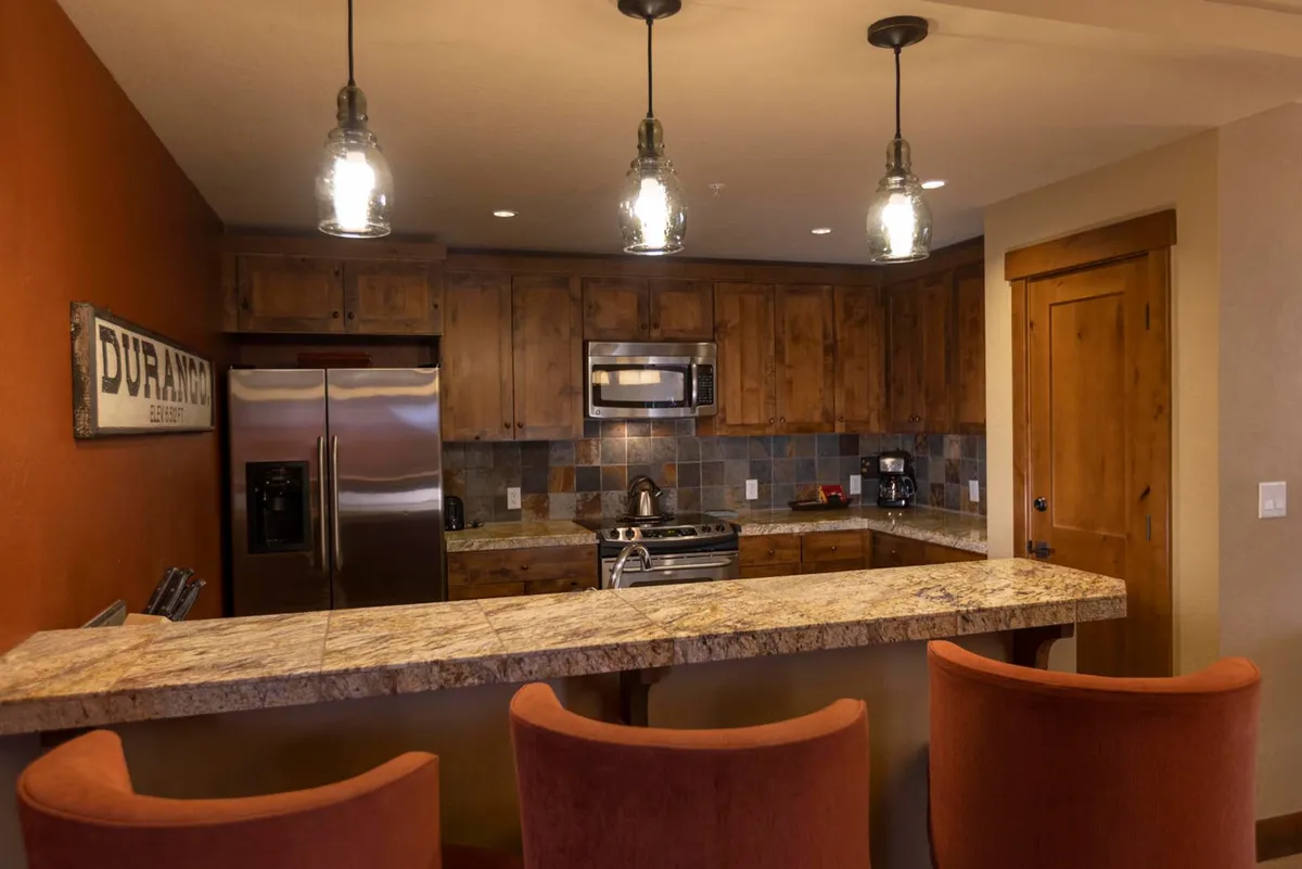 A modern kitchen with granite countertops, warm wooden cabinets, and sleek pendant lighting above the breakfast bar. Bar stools invite guests to linger, making it an ideal spot for cooking, casual dining, or late-night conversations. The kitchen's openness to adjacent living spaces is apparent.