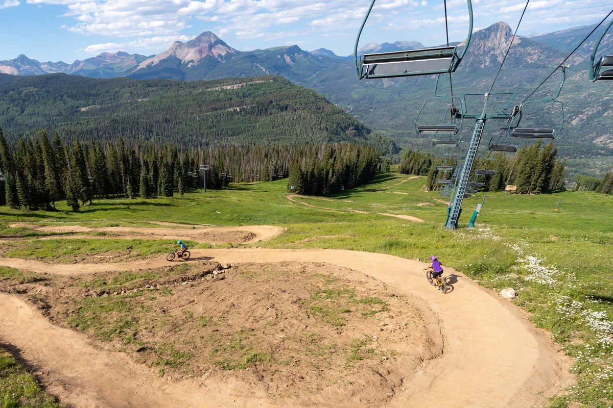 An inviting exterior scene showcasing mountain bike trails weaving through lush greenery, a ski lift poised for adventure, and the dramatic, snow-capped Needles Mountain Range rising in the distance. The vibrant landscape is bathed in natural light, evoking a sense of exhilaration and possibility.